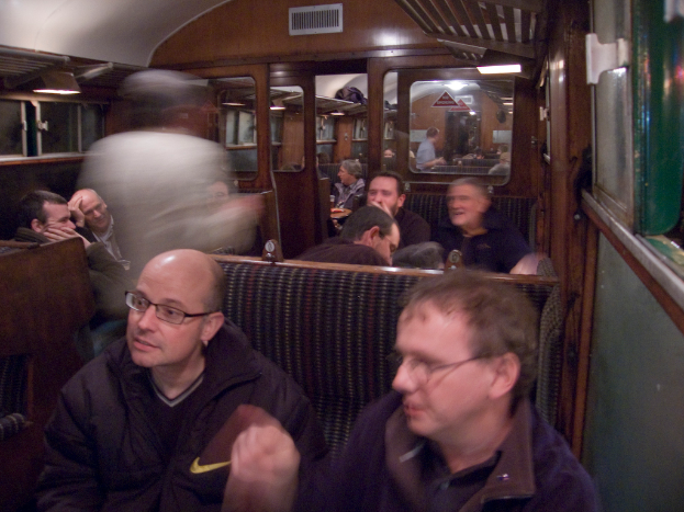 Zwei Männer sitzen auf einem Sitz in einem Abteil des Zugs, hinter ihnen ist eine Gruppe von Menschen zu sehen; oben ist ein Eingang zwischen den Abteilen zu sehen und rechts ein Fenster.