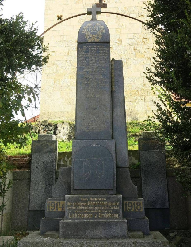 Ein Steindenkmal für deutsche Soldaten, die im Zweiten Weltkrieg starben, steht vor einer Kirche mit einem Kreuz, umgeben von Bäumen und einer Mauer.