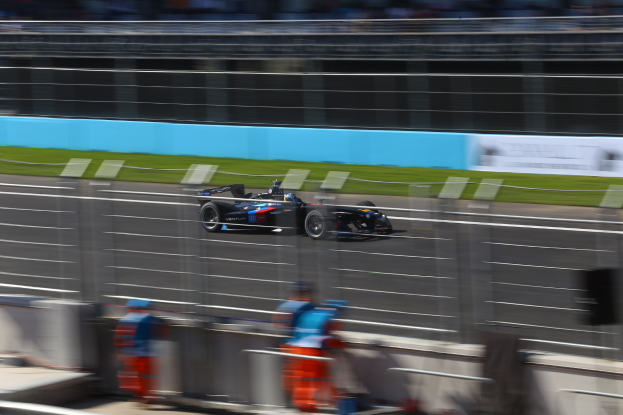 Ein Formel-1-Auto fährt auf einer Rennstrecke vorbei an einer Menge Zuschauern, Gras am Boden und Schildern im Hintergrund.