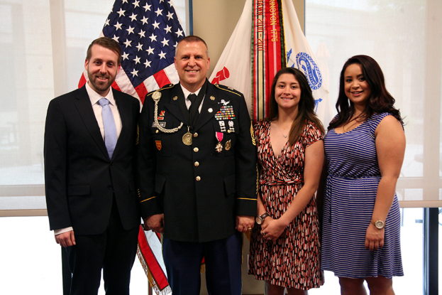 Zwei Frauen in Kleidern, eine Person in Uniform und ein Mann in einem Anzug stehen und lächeln drinnen, mit Jalousien und Flaggen im Hintergrund.
