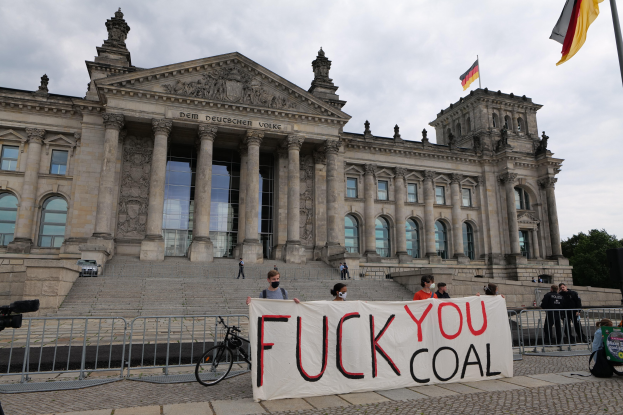 Eine Gruppe von Menschen mit einem Transparent mit der Aufschrift "Fuck You Coal" vor dem Reichstaggebäude in Berlin, Deutschland, mit sichtbaren architektonischen Details des Gebäudes, einem Fahrrad in der Nähe, Bäumen, einem Fahnenmast und einem bewölkten Himmel.