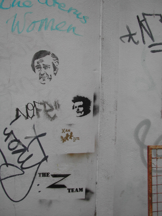 Ein verrosteter Zaun steht vor einer Wand, die mit Zeichnungen und Wörtern geschmückt ist.