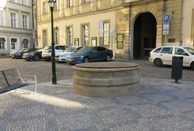 Ein Stadtplatz mit einer Bank in der Mitte, umgeben von Gebäuden mit Fenstern, Laternen und Schildern, mit einem zentralen Springbrunnen.