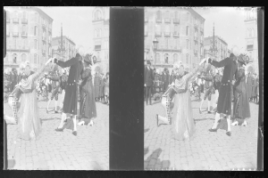 Ein Schwarz-Weiß-Foto einer Gruppe von Menschen, die auf der Straße tanzen, während eines Faschingsumzugs, mit Kostümen, umgeben von Gebäuden, Straßenlaternen und einer klaren Himmel.