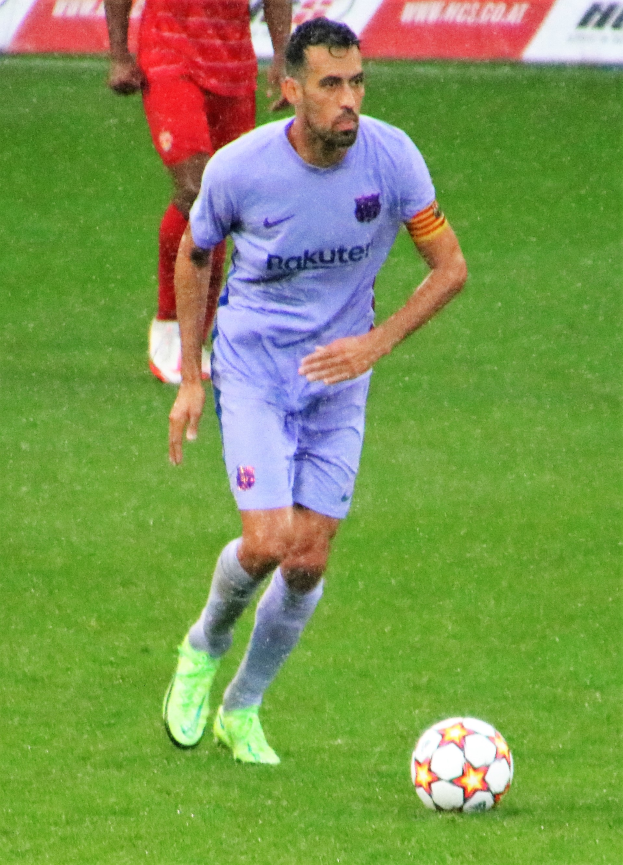 Ein Fußballspieler in einem Sportdress und -schuhen ist im Einsatz auf dem Feld, wobei ein Textschild im Hintergrund sichtbar ist.