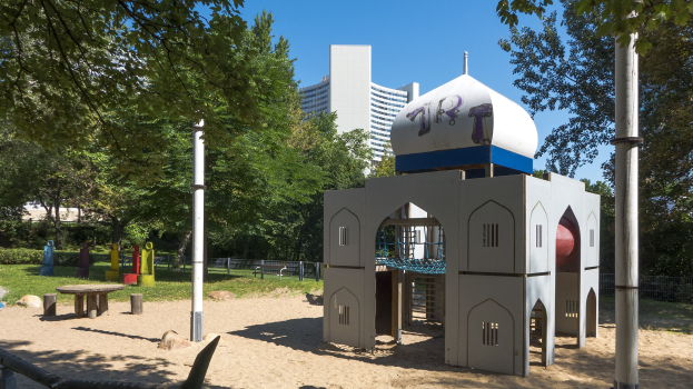 Ein Spielplatz mit einem zentralen Spielgerüst, umgeben von Bäumen, Pfählen, Bänken und Gebäuden unter einem klaren blauen Himmel.
