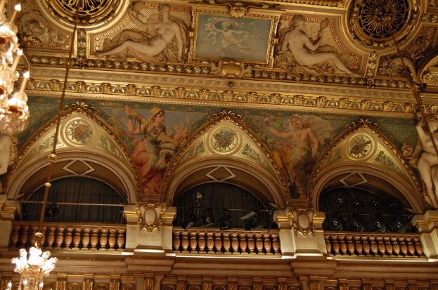Eine Wand mit Skulpturen, einem Bild und einem Rahmen, mit Kronleuchtern und Lichtern, einer Balkon mit Vorh√§ngen.