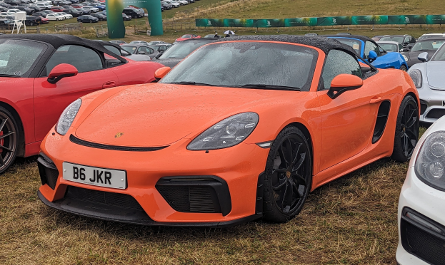 Porsche 718 Boxster GTS auf dem Goodwood Festival of Speed mit anderen Autos, einer Fahne, Bäumen und einem klaren blauen Himmel.