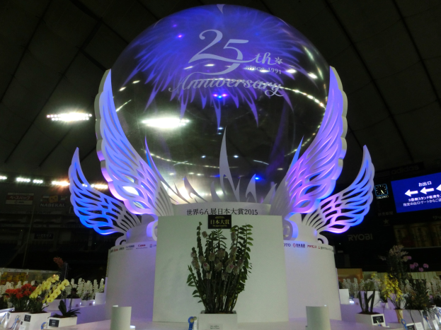 Ein großer Kristallball mit Flügeln oben drauf, umgeben von Blumenschmuck, Namensschildern und anderen Gegenständen auf dem Boden, vor einem Hintergrund aus Brettern, Lichtern und einem Bildschirm während einer 25-jährigen Jubiläumsfeier.