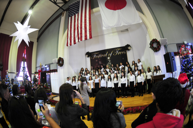 Gruppe von Menschen auf einer Bühne mit Weihnachtsdekoration, einem Rednerpult, Lautsprechern, einer Texttafel, Vorhängen, Flaggen und Deckenleuchten vor einem Publikum mit Kameras und Handys.