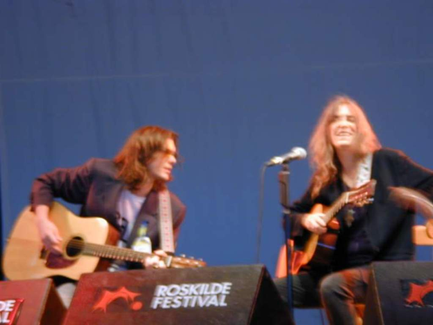 Zwei Männer auf der Bühne mit musikalischen Instrumenten, ein Mikrofon davor und zwei Boxen auf dem Boden mit "ROCK SLIDE FESTIVAL" darauf.