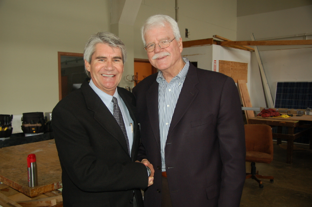 Zwei ältere Männer geben sich die Hand in einem Bürosetting mit einem Monitor, einem Tisch und einem Stuhl im Hintergrund.