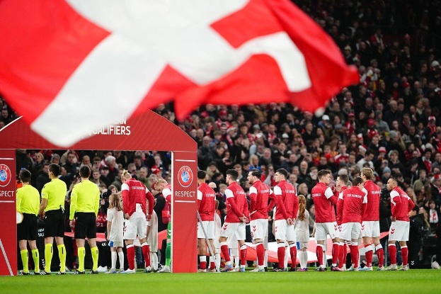 Gruppe von Menschen auf einem Fußballfeld mit einer roten und weißen Flagge im Vordergrund, einem Bogen mit 'Bayern München vs. Bayern München Wett-Tipps & Vorschau' Text im Hintergrund und einer großen Menschenmenge im Stadion.