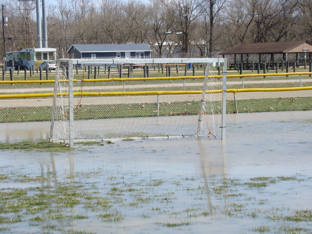 Fußballtor in einem überfluteten Feld mit umgebenem Gras und Wasser, Hütten, Pfosten, Bäumen, Fahrzeugen und einem klaren blauen Himmel im Hintergrund.
