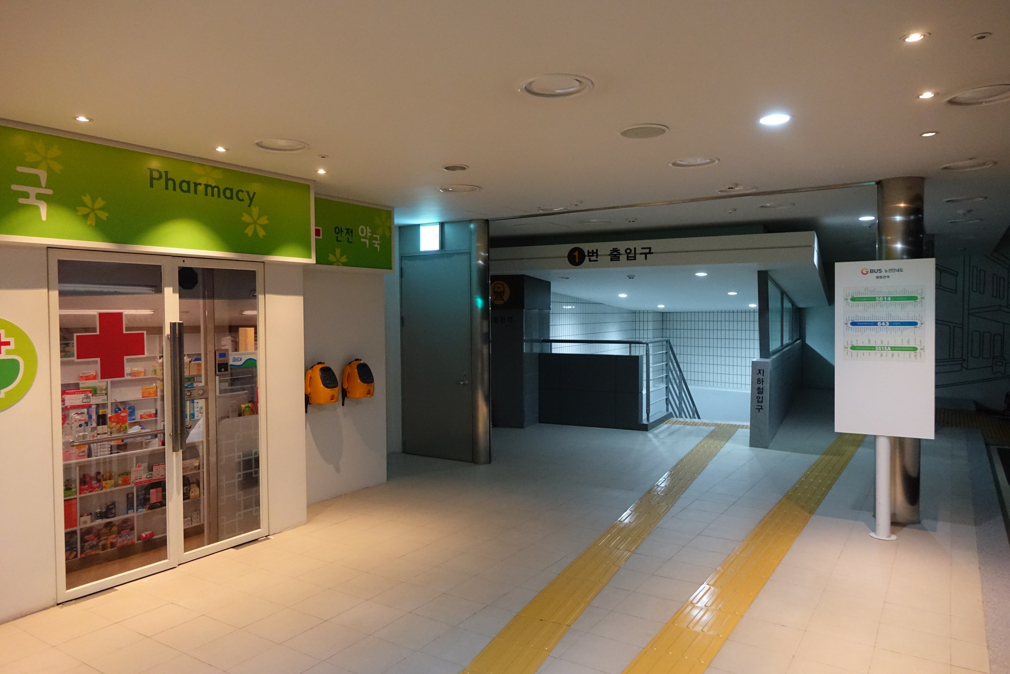 Apothekeneingang mit einem "Apotheke"-Schild auf der linken Seite, einer Tafel mit Text auf der rechten Seite, beleuchteten Lampen oben und unbestimmten Hintergrundgegenständen.