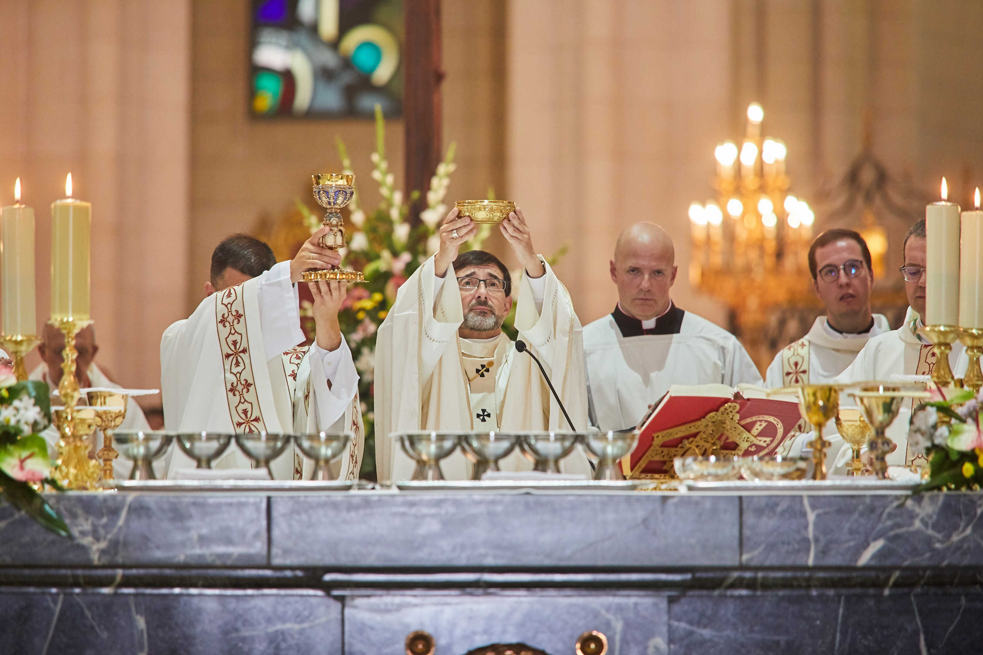 Eine Gruppe von Priestern vor einem Altar, jeder hält einen Kelch in der Hand, mit einem Tisch in der Mitte, auf dem Gläser, ein Mikrofon, ein Buch und andere Gegenstände stehen; ein Blumenstrauß links vom Altar und ein Kerzenständer rechts, mit einem Buntglasfenster im Hintergrund.
