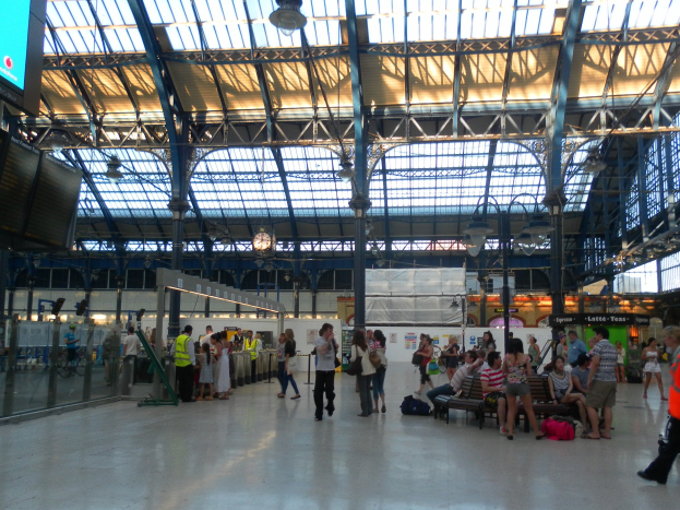 Menschen gehen und sitzen in einem belebten Bahnhof mit Taschen auf dem Boden, ein Anzeigebildschirm, Metallstangen, Deckenleuchten und eine Wanduhr.
