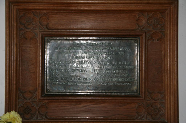 Eine Gedenktafel mit Text ist an einer braunen Wand oder einer hölzernen Tür angebracht, mit einer gelben Blumenvase in der linken unteren Ecke.