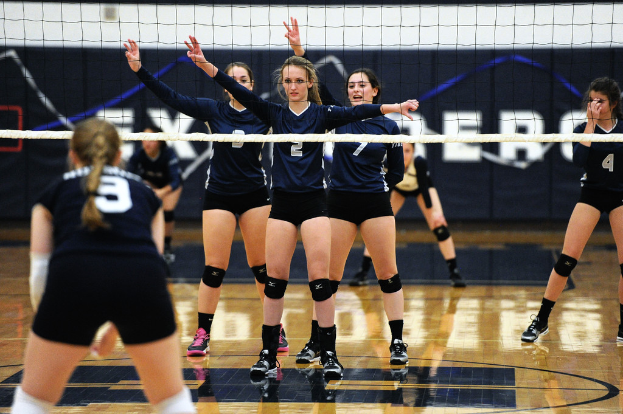 Drei Mädchen bei einem Volleyballspiel auf dem Feld stehend, die Hände erhoben, ein Netz davor und ein Zaun im Hintergrund.