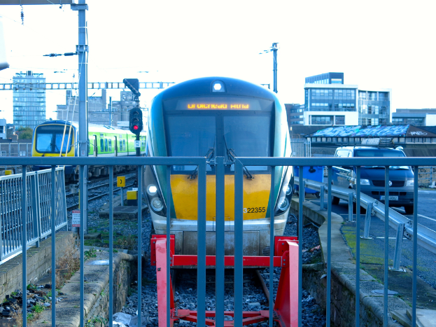 Zwei Züge auf dem Gleis mit einem Zaun im Vordergrund, Masten auf dem Land, einer mit einer Signallampe, ein Auto auf der Straße rechts, Gebäude im Hintergrund und der Himmel darüber.