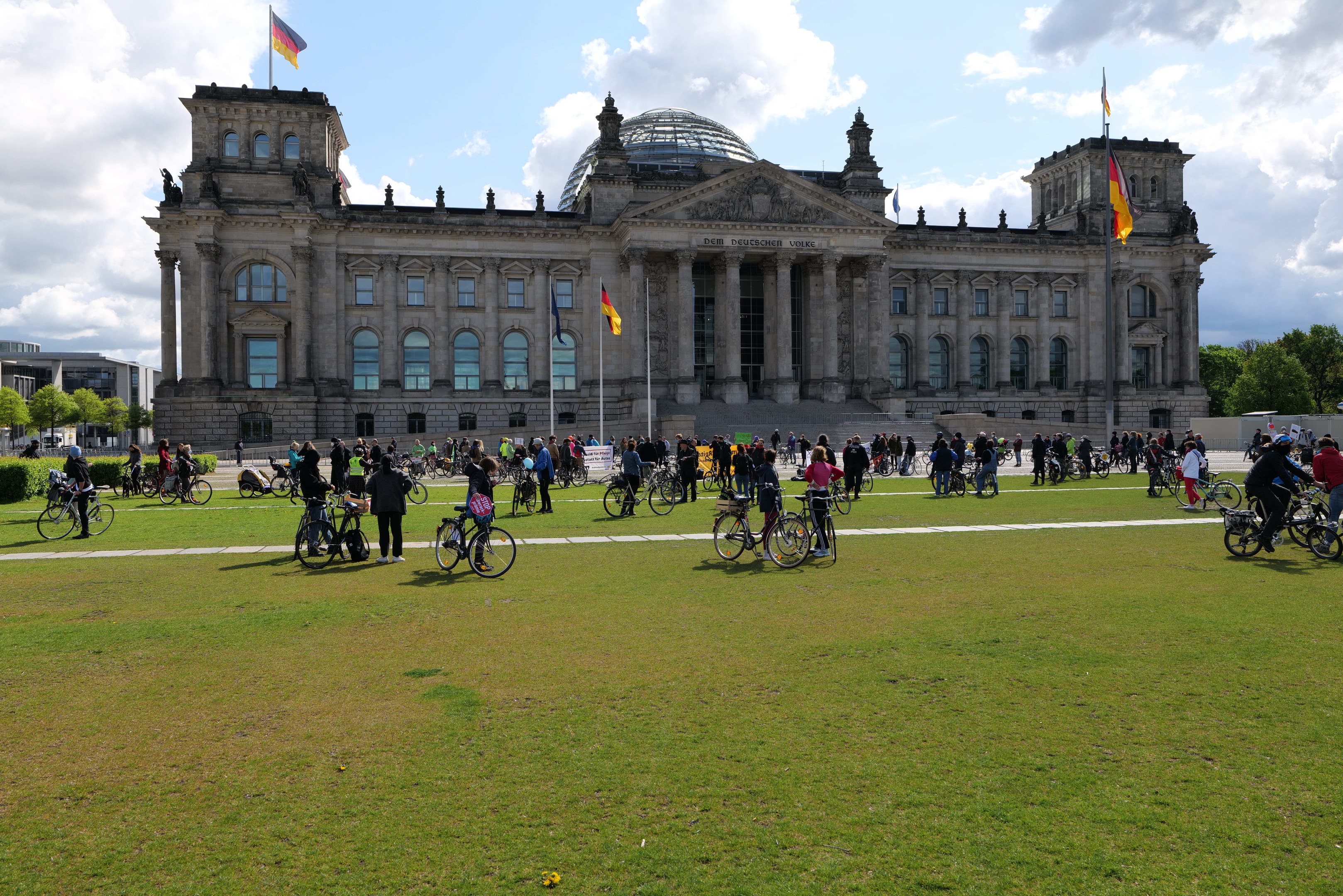 Eine Gruppe von Menschen, die vor dem Reichstaggebäude in Berlin, Deutschland, Fahrräder fährt, mit Bäumen, Pflanzen und einem bewölkten Himmel.