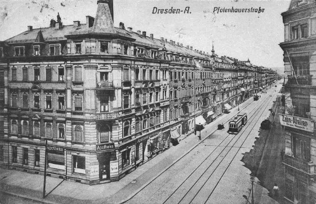 Ein altes Schwarz-Weiß-Foto einer Stadtstraße in Dresden, Deutschland, mit Fahrzeugen, Fußgängern, Gebäuden mit Fenstern und Schildern, Strommasten mit Drähten und einem Himmel im Hintergrund, mit Text oben und unten auf dem Bild.