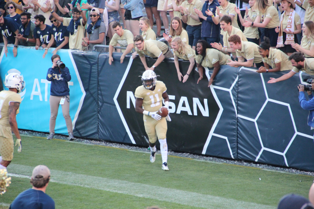 Ein Fußballspieler läuft mit dem Ball auf einem Rasenfeld vor einer Menge, einige halten Kameras und tragen Helme, mit einem Banner im Hintergrund.