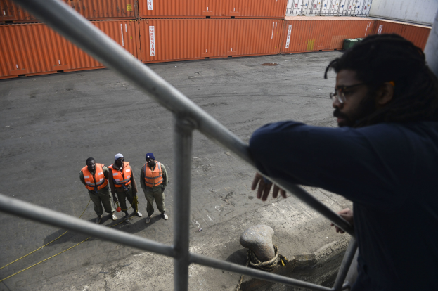 Ein Mann in legerer Kleidung steht auf einem Metallgeländer in der Nähe einer Gruppe von Menschen mit orangen Jacken und Kappen, mit Containern und einer Wand im Hintergrund am Hafen von Los Angeles.