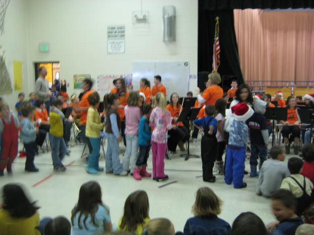 Kinder in bunten Kleidern sitzen unten im Bild, mit einer Bühne, einem Vorhang, einer weißen Tafel, Plakaten, einer Tür, Schildern, einer Fahne und einem Objekt an der Wand im Hintergrund.