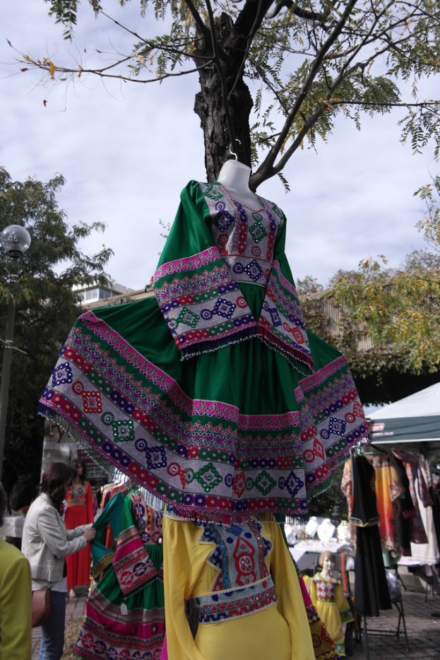 Eine Schaufensterpuppe steht in der Mitte eines belebten Einkaufsgebiets mit mehreren Geschäften, einer Person, die Kleider an einer Stange arrangiert, einem Gebäude und Bäumen im Hintergrund.