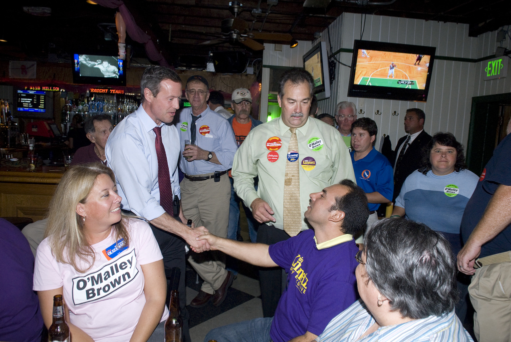 Drei Personen sitzen unten auf dem Bild, mit zwei Glasflaschen vor ihnen; der Mann im weißen Hemd gibt dem Mann im blauen T-Shirt die Hand, während viele Menschen hinter ihnen stehen. Links steht ein Tisch mit einigen Gegenständen, und im Hintergrund zeigt ein Fernsehbildschirm Inhalt. Rechts ist eine Ausgangstafel neben einer weißen Wand mit Fernsehgeräten zu sehen.