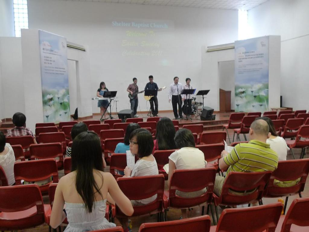 Eine Buhnenböhne mit zwei Menschen, die in der Mitte Gitarre spielen, und anderen, die in den Ecken singen und Instrumente spielen, mit Bannern neben der Bühne und einem Publikum davor, das Papiere und Büher hält.