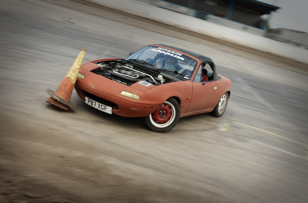 Eine Person in einem roten Auto auf einer Rennstrecke, die auf eine Verkehrsinsel zusteuert, mit einem unscharfen Hintergrund, der hohe Geschwindigkeit andeutet.