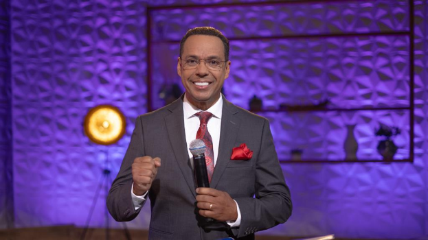 Stephen Colbert in Anzug, Krawatte und Brille auf einer Bühne mit Mikrofon stehend, mit einer Wand und verschiedenen Gegenständen im Hintergrund.