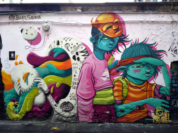 Ein buntes Wandgemälde an der Seite eines Gebäudes, teilweise von Graffiti bedeckt, mit einer Straße im Vordergrund, Strommasten und einem klaren blauen Himmel im Hintergrund.