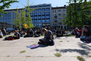 Menschen sitzen vor einem Gebäude auf dem Boden während einer Demonstration in Berlin, einige tragen Masken, mit verstreuten Taschen und Gegenständen, unter einem klaren blauen Himmel.
