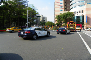 Ein Polizeiwagen fährt auf einer Straße mit hohen Gebäuden, Bäumen, Laternen, Ampeln und Passanten unter einem bewölkten Himmel.