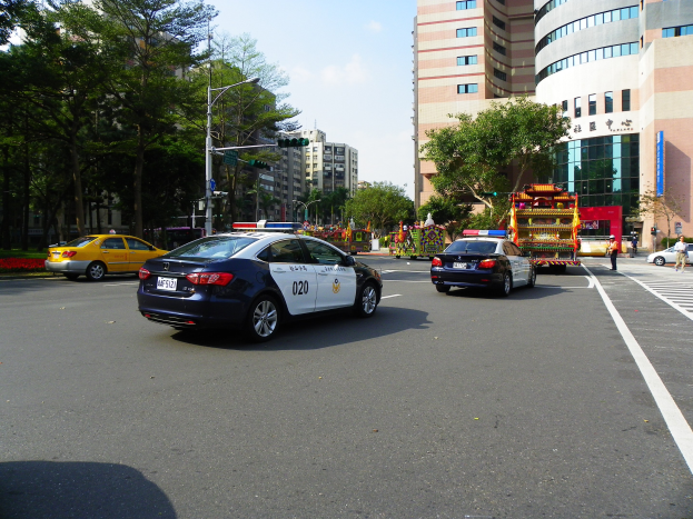 Ein Polizeiwagen fährt auf einer Straße mit hohen Gebäuden, Bäumen, Laternen, Ampeln und Passanten unter einem bewölkten Himmel.