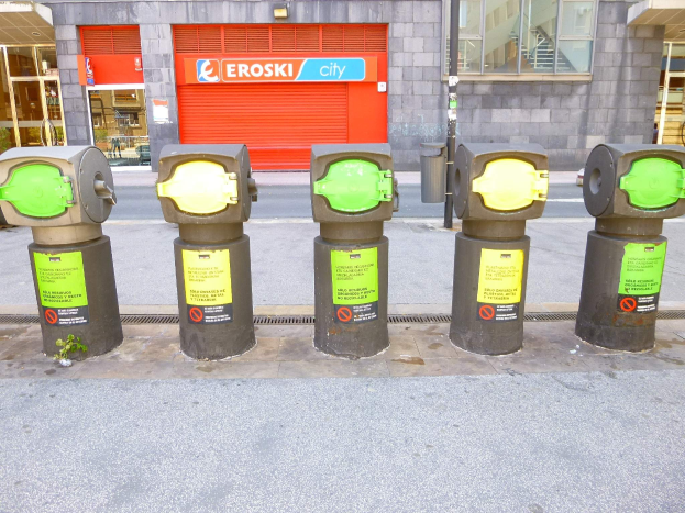 Eine Reihe von Parkautomaten entlang einer Straße, mit einem Gebäude mit Glasfenstern und einem Rollladen mit Text im Hintergrund und einem näheren Müllcontainer.