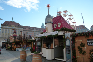 Ein lebendiger Weihnachtsmarkt in einem Stadtplatz, umgeben von Gebäuden, Straßenlaternen und geschmückten Bäumen, mit Menschen, die gehen und Fahrzeugen auf der Straße unter einem bewölkten Himmel.