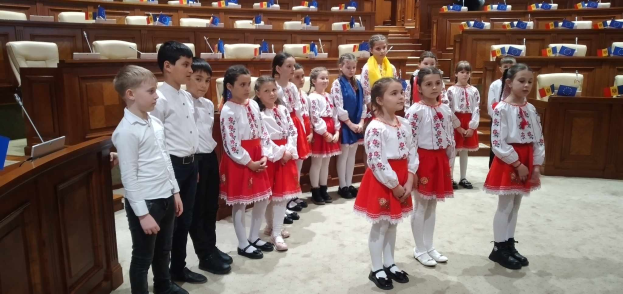 Gruppe von Kindern in weißen und roten Käppchen, die zusammen in einem Raum mit einem Podium, Mikrofon, Stühlen, Tischen und Fahnen bei einer Nationalversammlung stehen.