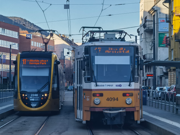Zwei gelbe und weiße Straßenbahnen auf einer Stadtstraße mit hohen Gebäuden, Fahrzeugen, Geländern, Polen, Schildern, Bäumen, einem Hügel und dem Himmel im Hintergrund.
