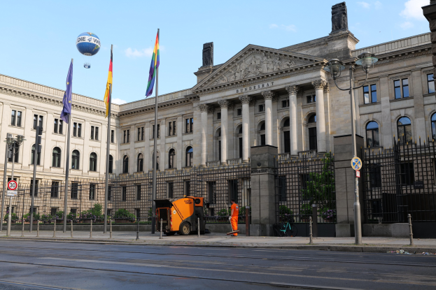 Großes modernes Regierungsgebäude mit vielen Fenstern, umgeben von Straßeninfrastruktur, Fußgängern und Bäumen, mit bewölktem Himmel im Hintergrund.