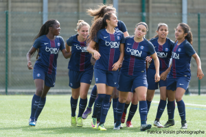 Eine Gruppe junger Frauen, die auf einem Rasenfußball spielen, mit einem Maschendrahtzaun und Pfosten im Hintergrund und einer Wand dahinter, mit dem Text "Paris Saint-Germain Frauenfußball" in der rechten unteren Ecke.