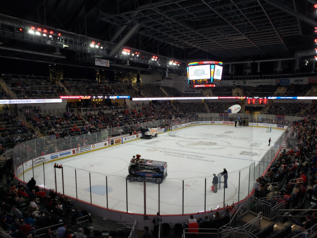 Ein Eishockeyspiel in einer großen Arena mit Fahrzeugen auf dem Eis, Zuschauern um die Bahn und Menschen auf den Tribünen, mit Wänden, Lichtern und einem Bildschirm im Hintergrund.