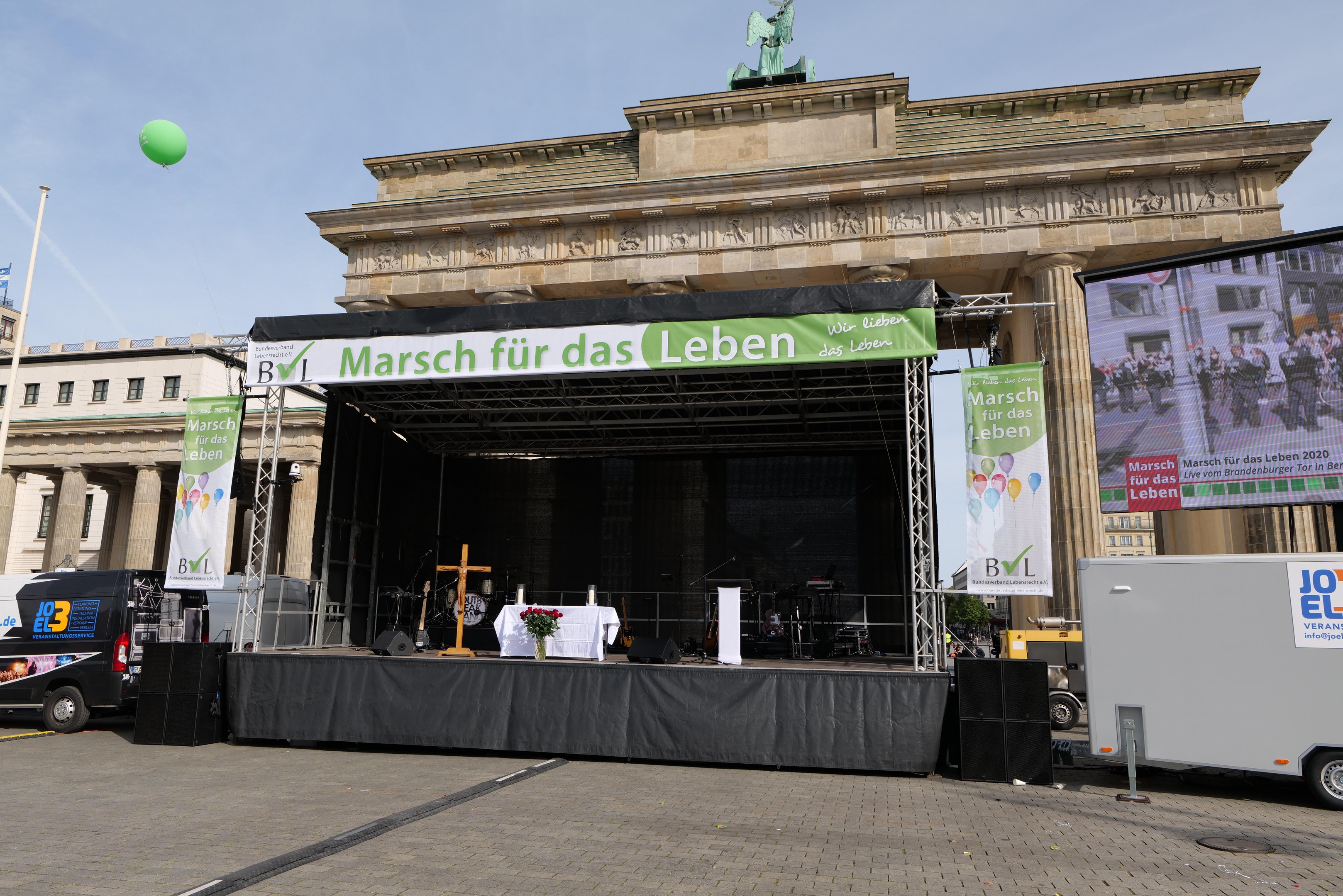 Bühne vor dem Brandenburger Tor in Berlin, Deutschland, mit einem Tisch mit einer Blumenvase, einem Kreuzsymbol, Bannern mit Text, Lautsprechern und anderen Gegenständen, sowie Fahrzeugen auf der Straße und Gebäuden im Hintergrund.
