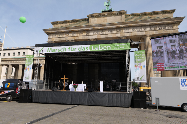 Bühne vor dem Brandenburger Tor in Berlin, Deutschland, mit einem Tisch mit einer Blumenvase, einem Kreuzsymbol, Bannern mit Text, Lautsprechern und anderen Gegenständen, sowie Fahrzeugen auf der Straße und Gebäuden im Hintergrund.