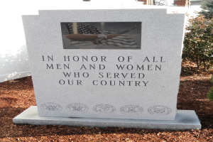 Ein Gedenkstein mit der Inschrift "In Ehren aller Männer und Frauen, die unserem Land gedient haben", flankiert von Pflanzen auf der rechten Seite und einem Gebäude im Hintergrund.