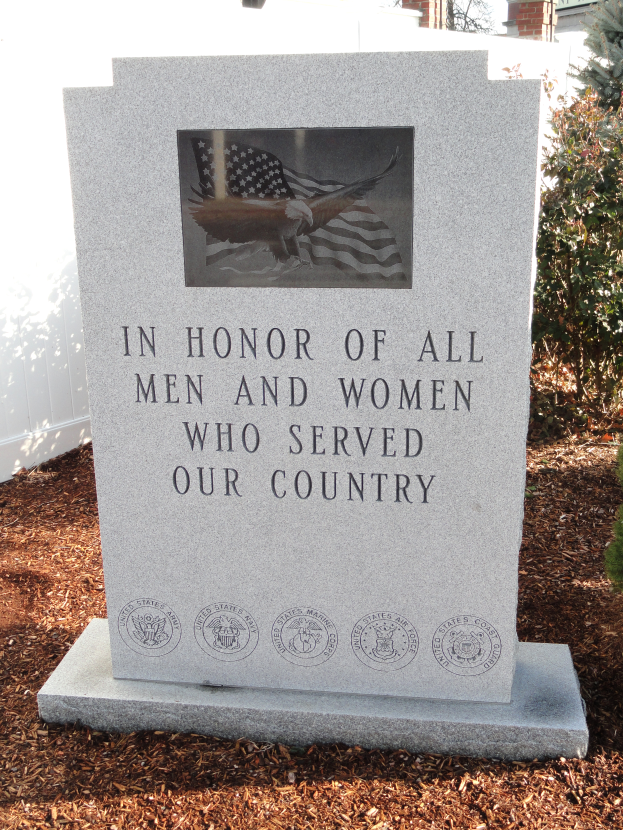 Ein Gedenkstein mit der Inschrift "In Ehren aller Männer und Frauen, die unserem Land gedient haben", flankiert von Pflanzen auf der rechten Seite und einem Gebäude im Hintergrund.