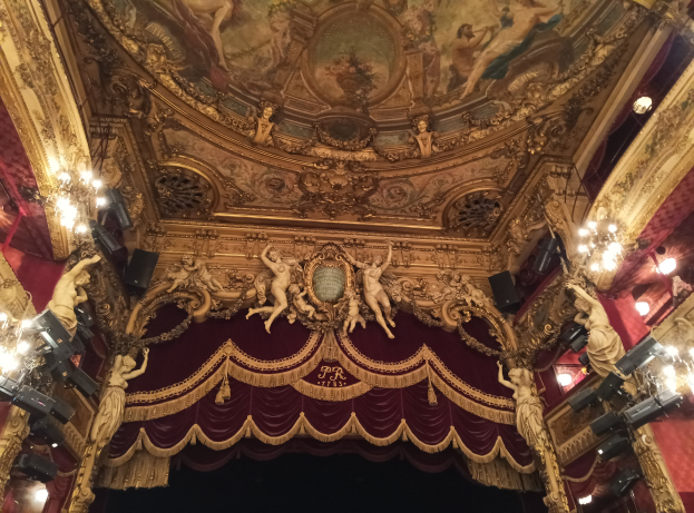 Innere eines Theaters mit rotem Vorhang, Kronleuchtern, Skulpturen, Lichtern und einer bemalten Decke, mit einer Bühne und ein paar Menschen unten.